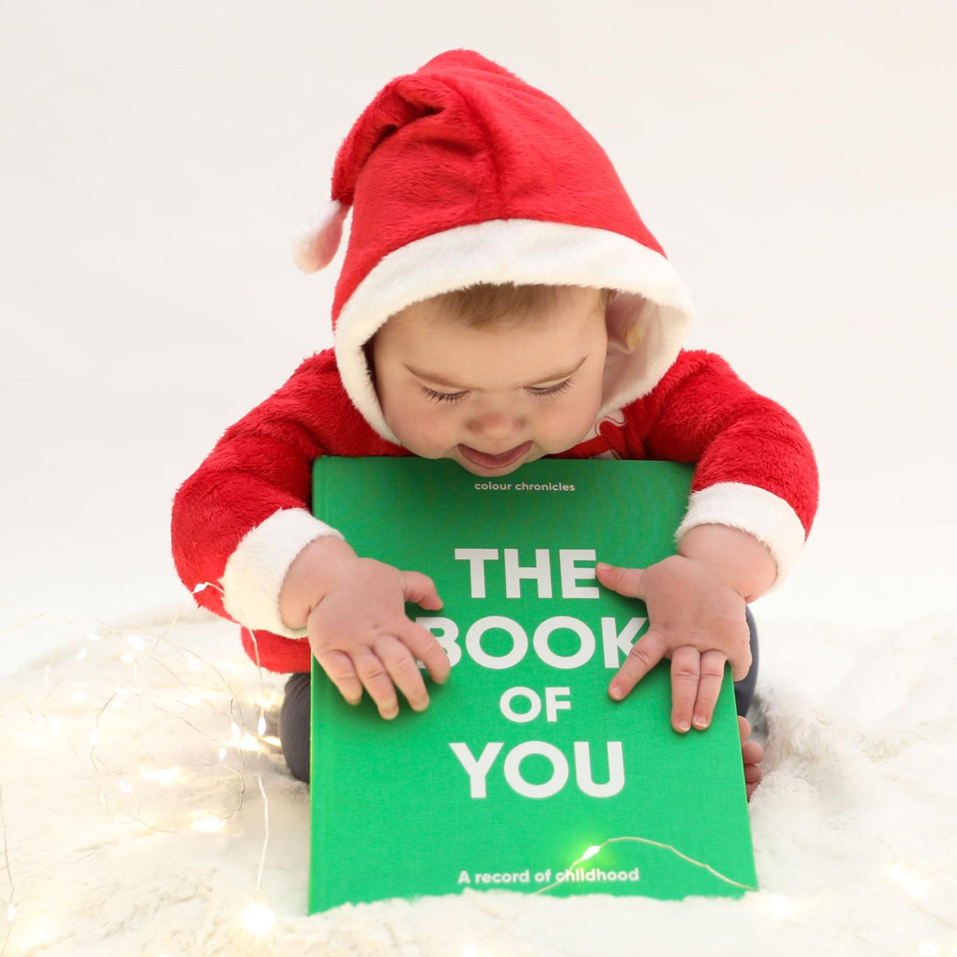 Baby in a Christmas Santa outfit holding a baby memory book, The Book of You by Colour Chronicles.