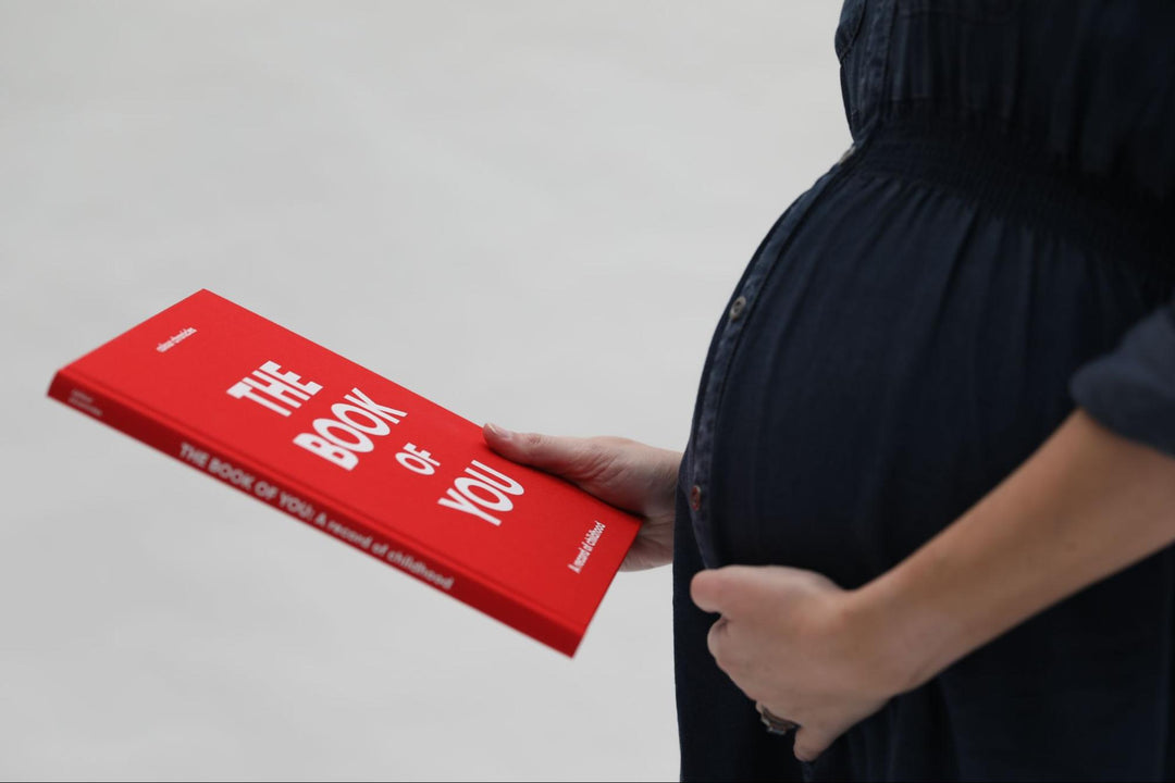 Baby mother holds her bump while looking at the baby memory book they’ve received as a gift, The Book of You by Colour Chronicles. 